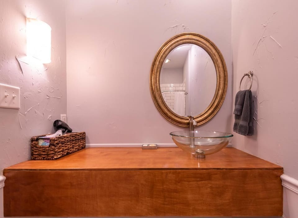 Bathroom vanity with round gold-framed mirror, glass bowl sink, wooden countertop, and textured light walls.