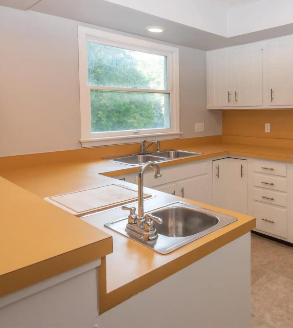 Kitchen with mustard-yellow laminate countertops, white cabinetry, dual stainless steel sinks, and a large window.
