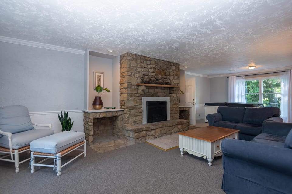 Living room with blue upholstered sofas, stone fireplace, white wood coffee table, and cushioned armchair with ottoman.