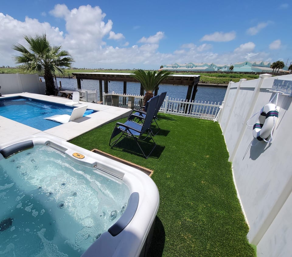 Hot tub bubbles beside a rectangular concrete pool, blue reclining chairs on artificial turf near waterway dock.