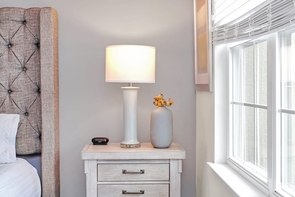 A bedside table with a modern lamp and decorative vase next to a window with white blinds.