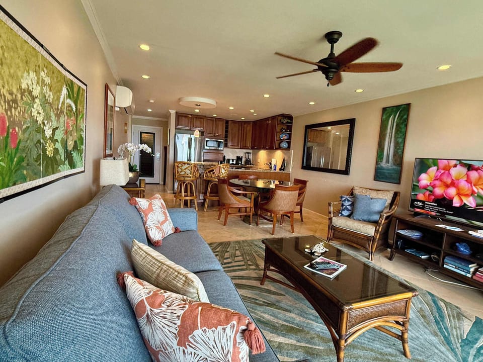 Spacious Living Area – Grey Sofa with Floral Pillows, Wicker Armchair, Flat-Screen TV, Dining Area, and Open Kitchen with Modern Appliances.