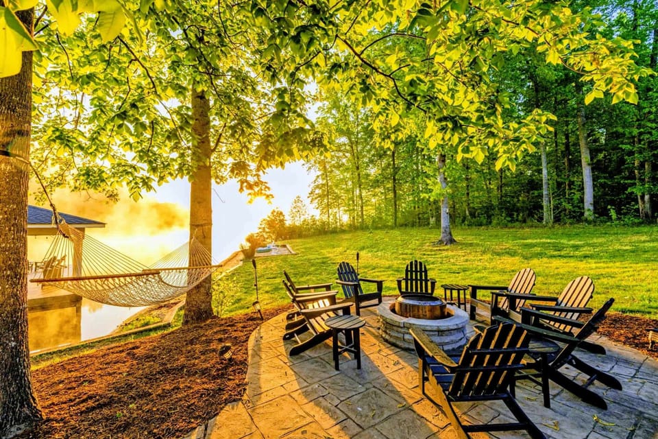 A stamped-concrete fire pit patio with Adirondack seating around a stone fire ring and a lakeside hammock catching the golden hour light through the trees.