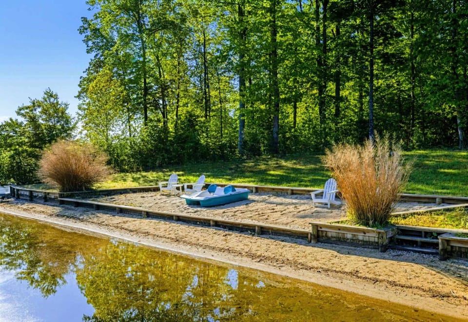 A private sandy beach with Adirondack seating, a paddleboat at the waterline, and ornamental grasses framing the cove views.