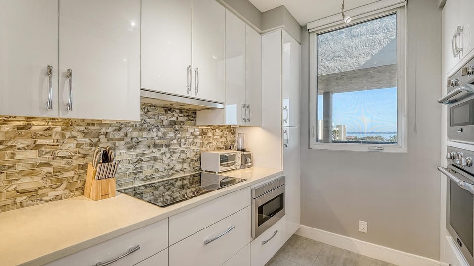 Galley kitchen with glossy white cabinetry, marbled tile backsplash, electric cooktop, and window with partial water view.