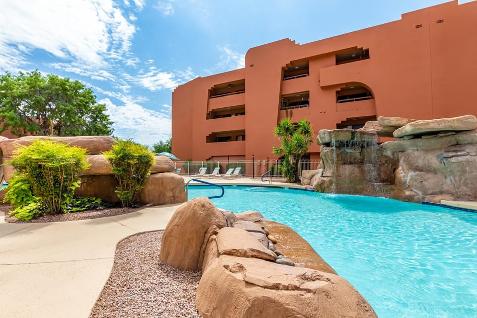 Outdoor pool area with rock waterfall feature, lounge chairs, desert landscaping, and terracotta building facade.