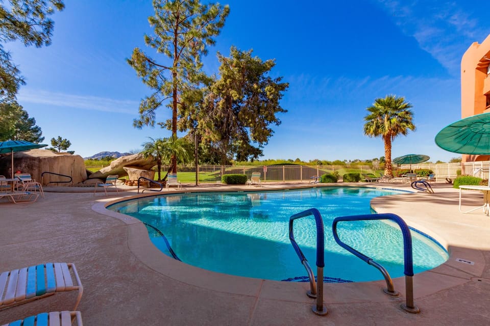 Curved pool edged by beige concrete deck with lounge chairs and green umbrellas under clear blue sky.