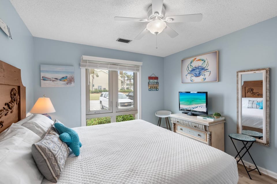 Bright coastal guest bedroom with a comfy king bed, beach-themed decor, flat-screen TV, and natural light pouring in through large windows—your perfect retreat after a day in the sun.