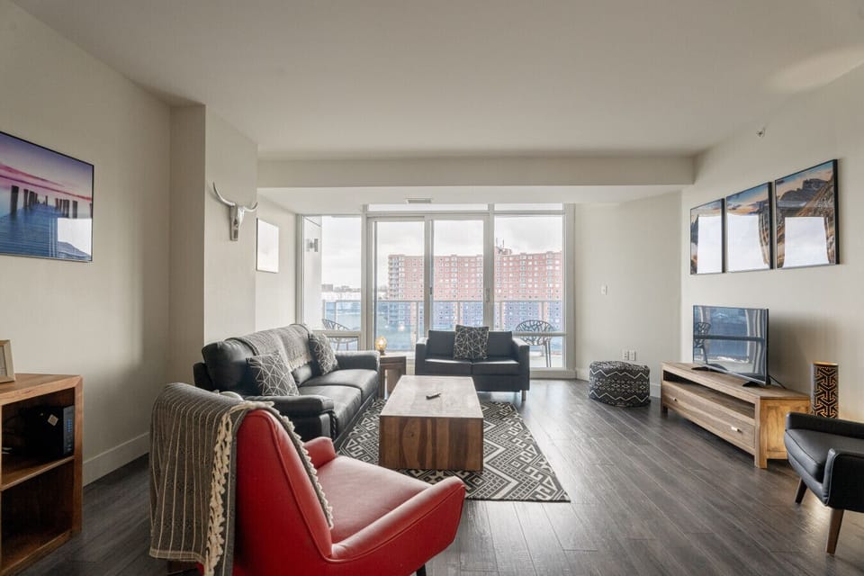 Welcome to your bright and inviting living area, the heart of this stylish apartment. Bathed in natural light from the massive glass doors, this space is designed for relaxation. Whether you’re curling up on the plush black sofa, reading in the striking r