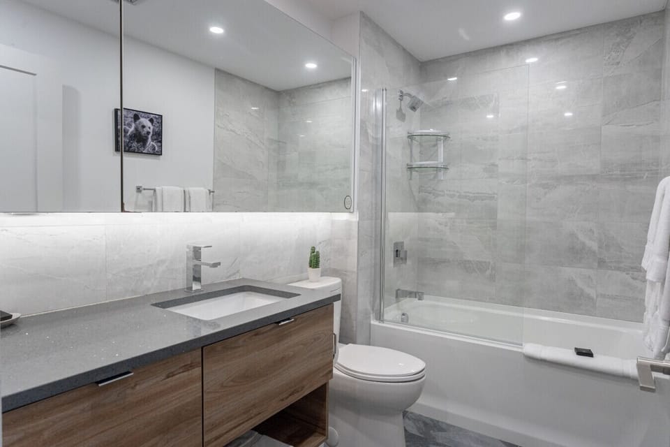 `Modern full bathroom featuring a floating wooden vanity with dark quartz countertop, under-mirror lighting, glass-enclosed tub/shower combination, and sleek grey wall tiles.`