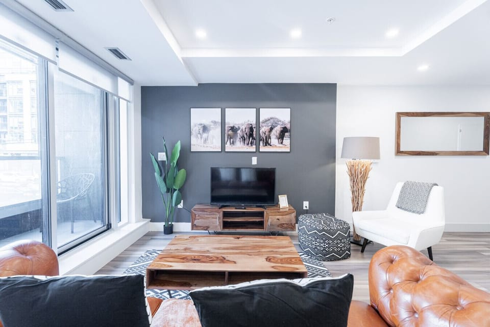 `Modern and inviting living room featuring a dark accent wall with elephant triptych art, a flat-screen TV on a wooden console, a plush white armchair, and a solid wood coffee table.`