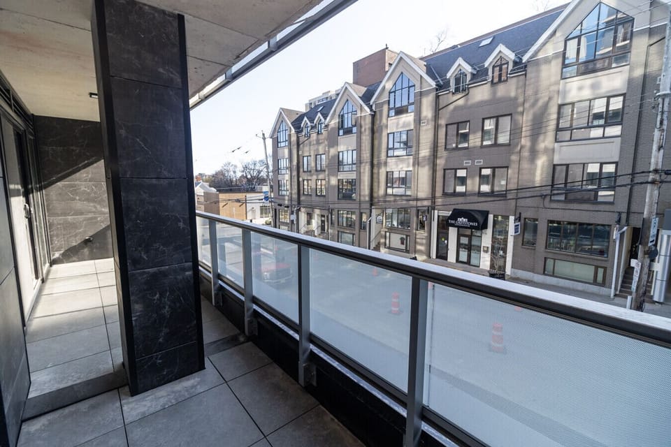 `Covered private balcony featuring dark accent pillars, glass railings, tile flooring, and street views of neighboring architecture.`