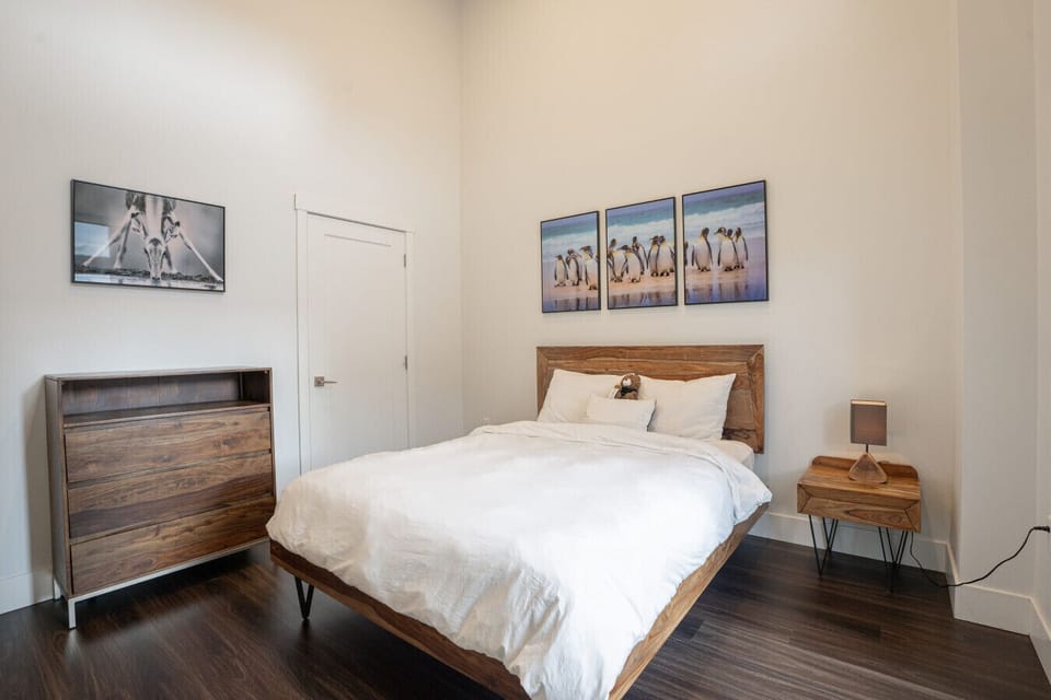 Spacious secondary bedroom featuring a mid-century modern wooden bed frame, plush white bedding, matching wood dresser and nightstand, crisp neutral walls, and high soaring ceilings for an open, airy feel.