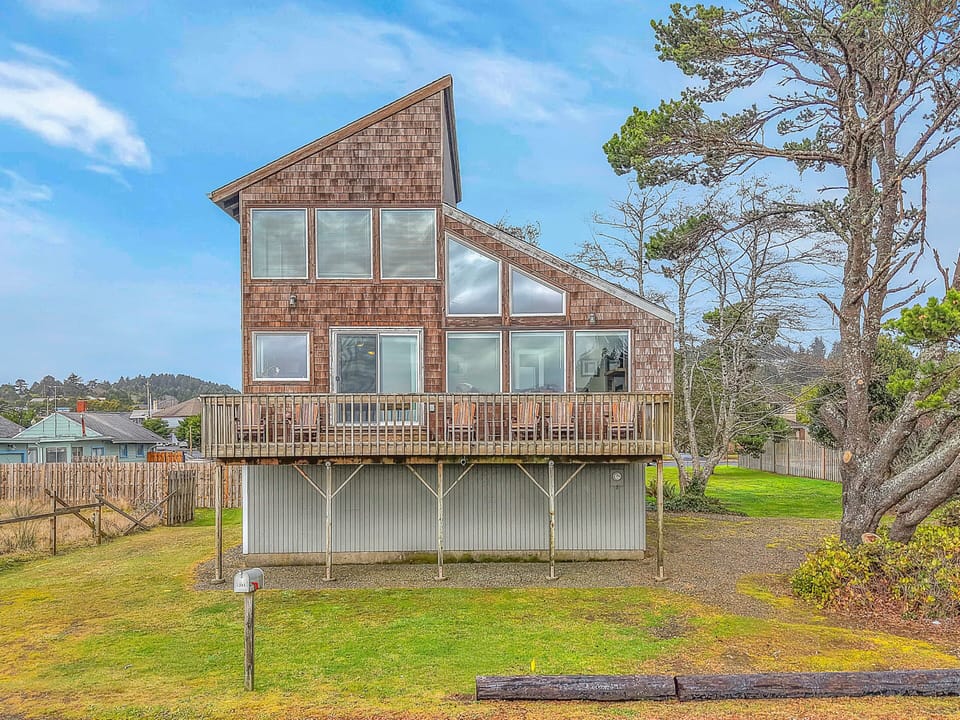 Contemporary cedar-shingled retreat elevated among coastal trees, featuring expansive windows and wraparound deck in peaceful neighborhood setting.
