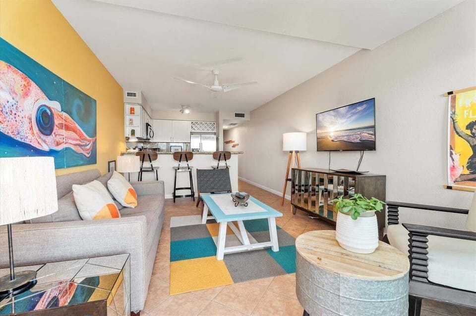 Beach condo living area with sofa, TV, coffee table, ceiling fan, and open kitchen view.