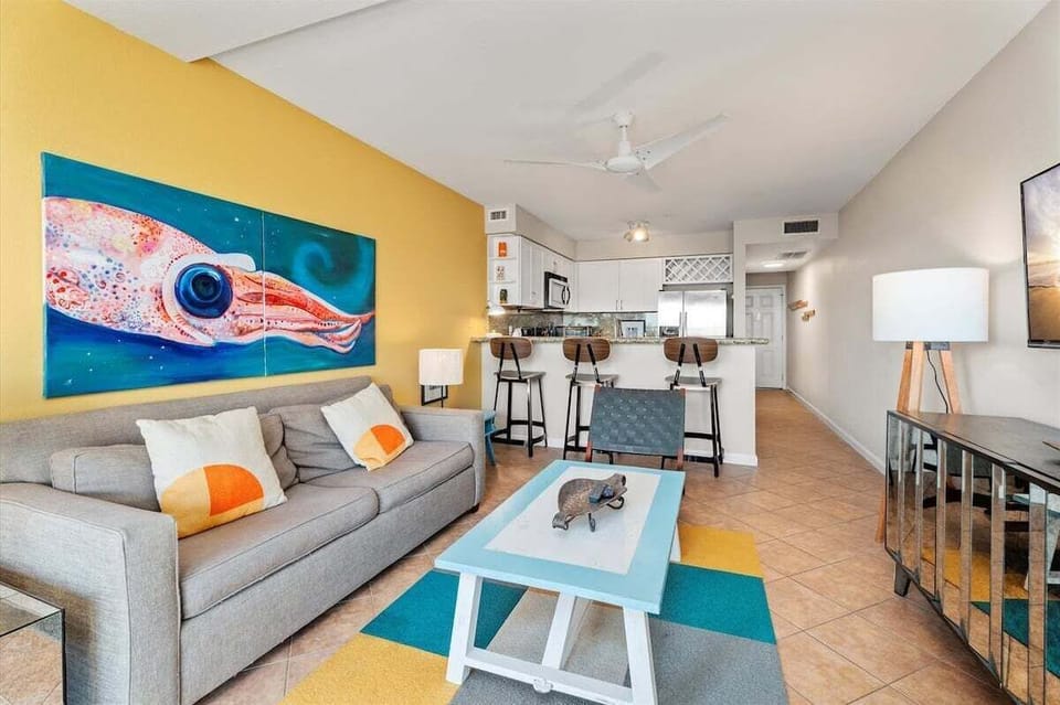 Open living room with sofa, colorful decor, ceiling fan, and kitchen breakfast bar.