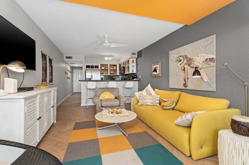 Open living room with yellow sofa, dining area, kitchen bar seating, TV, and beachy Galveston condo decor.
