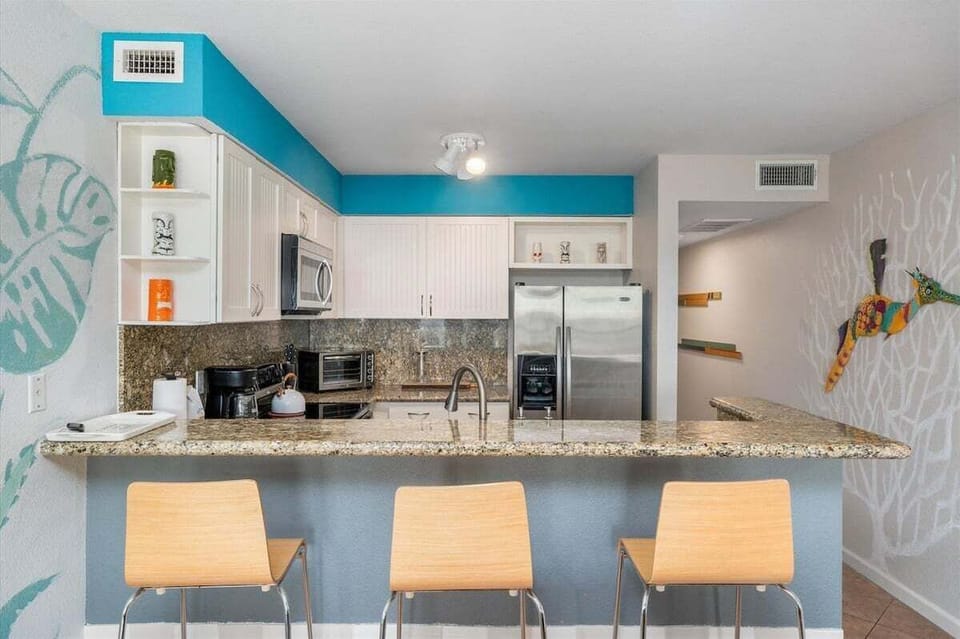 Fully equipped kitchen with granite countertops, stainless steel refrigerator, microwave, breakfast bar seating, and ample cabinet storage.
