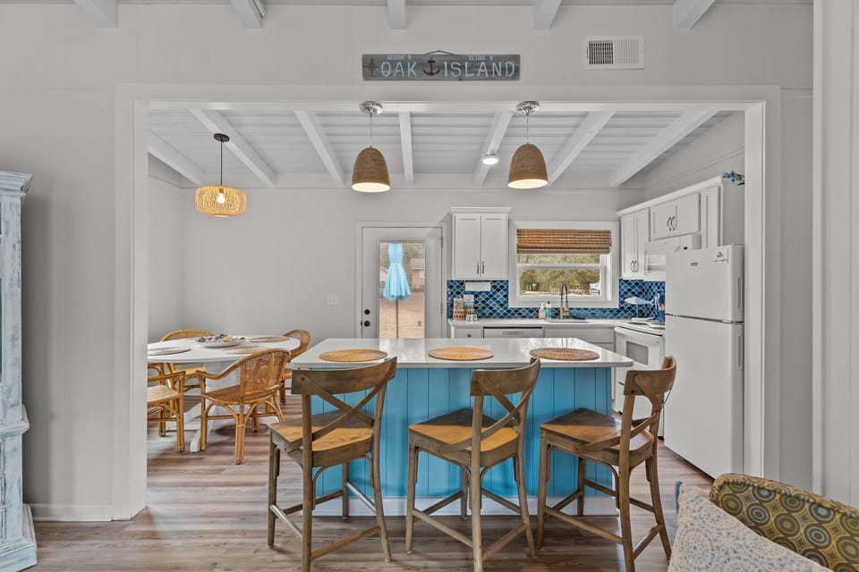 Bright open kitchen and dining area in Oak Island NC featuring a large blue island with seating, coastal lighting, round dining table with wicker chairs, and views into the comfortable living room.