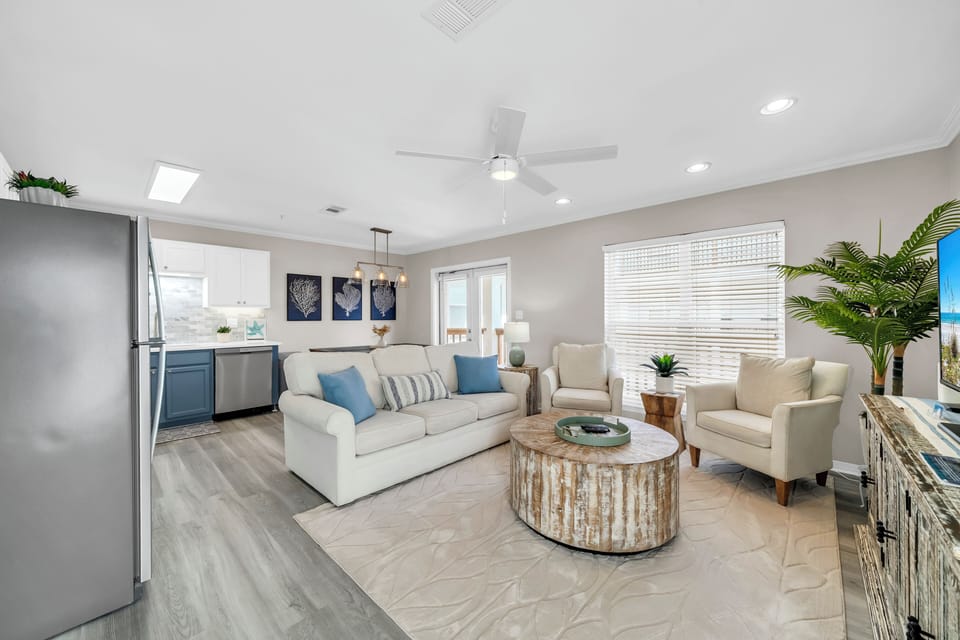 Sink into the plush coastal sofa and unwind in your bright, breezy living space featuring soothing beach-inspired decor, modern kitchen access, and natural light streaming through plantation shutters.
