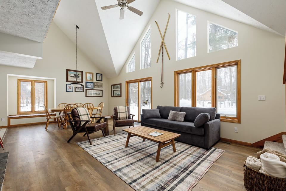 Soaring ceilings & expansive windows fill the living room w/ natural light, anchored by cozy seating & warm wood accents.