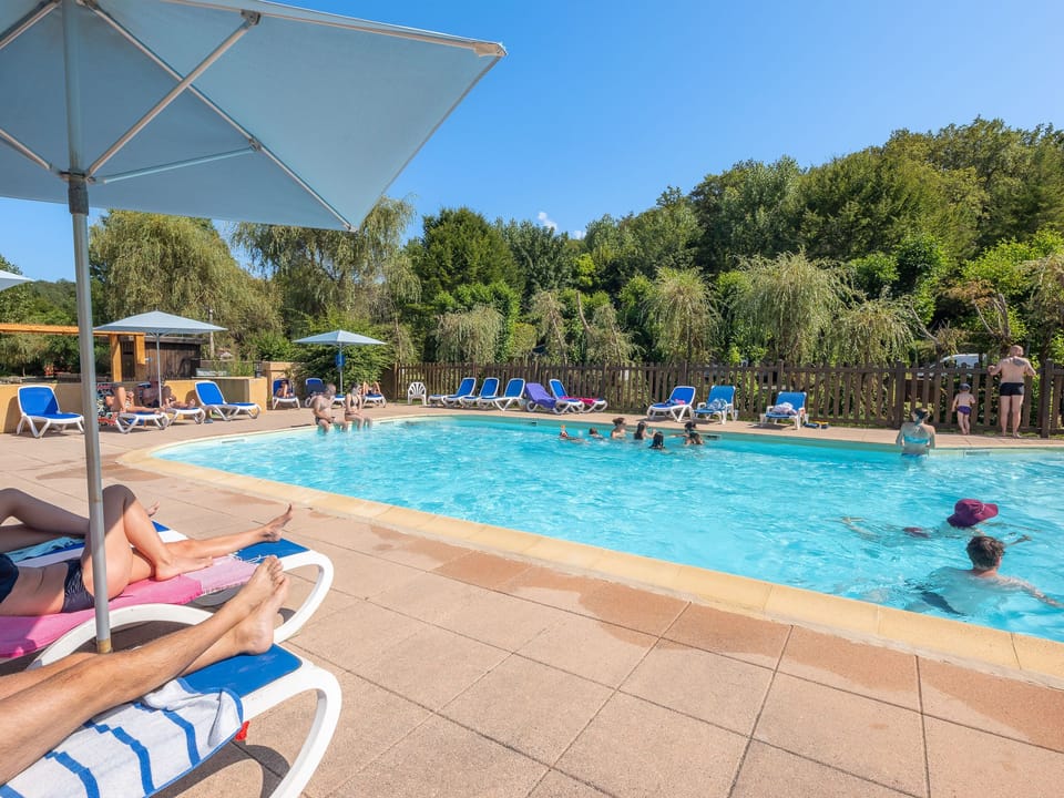 Sunbathers relax under blue umbrellas by the pool, creating a serene atmosphere for families to unwind in nature.