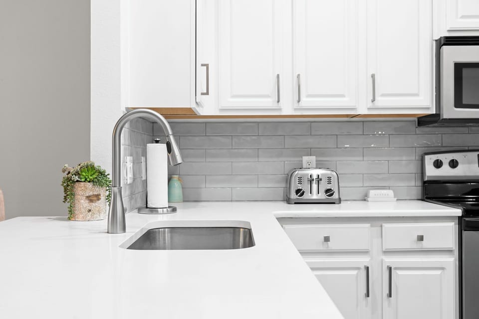 - Sleek countertops and a graceful gooseneck faucet make meal prep a breeze
- Stainless steel toaster and charming butter dish bring function and personality
- Glossy subway tiles and beautiful greenery complete the clean, coastal look