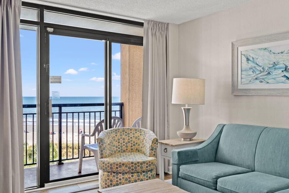 Sunlit one-bedroom living room featuring panoramic floor-to-ceiling windows with sweeping ocean and resort views. Coastal furnishings, open layout, and warm natural light create a serene and inviting Myrtle Beach getaway atmosphere.