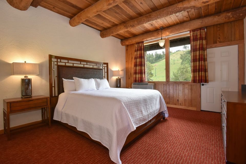 Rustic bedroom with exposed wooden beams, a plush bed, and large windows offering serene views of the outdoors.