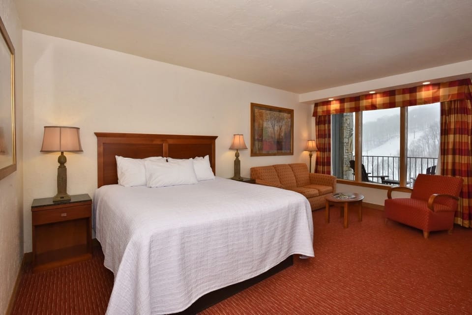 Spacious bedroom with a plush bed, cozy seating area, and large windows offering natural light and scenic balcony views.