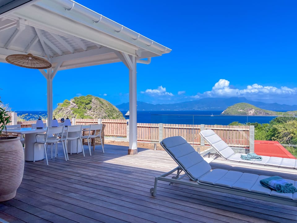 A wooden terrace with comfortable sun loungers and a dining table under a canopy, offering a stunning ocean view.