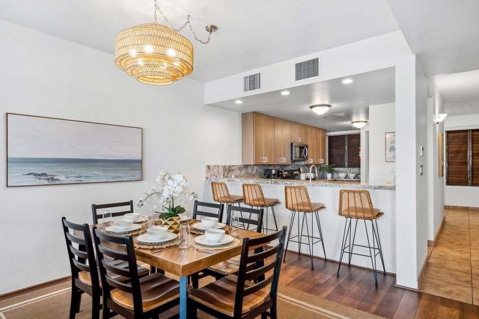 -Alternate dining area with wooden table and woven chandelier, kitchen behind with granite counters and wicker bar stools.