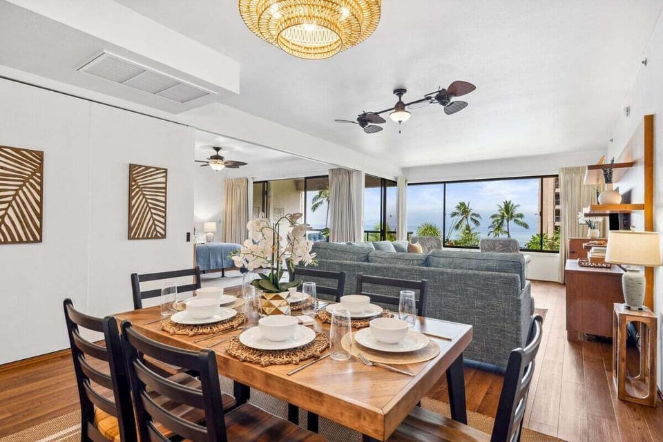 Dining table set for six with white plates and floral centerpiece, windows opening to palm trees and ocean views.