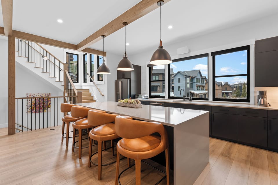Gather around the sleek island where natural light floods through expansive windows, highlighting the warm leather seating and elegant wood beams overhead—your perfect spot for morning coffee or evening conversation.