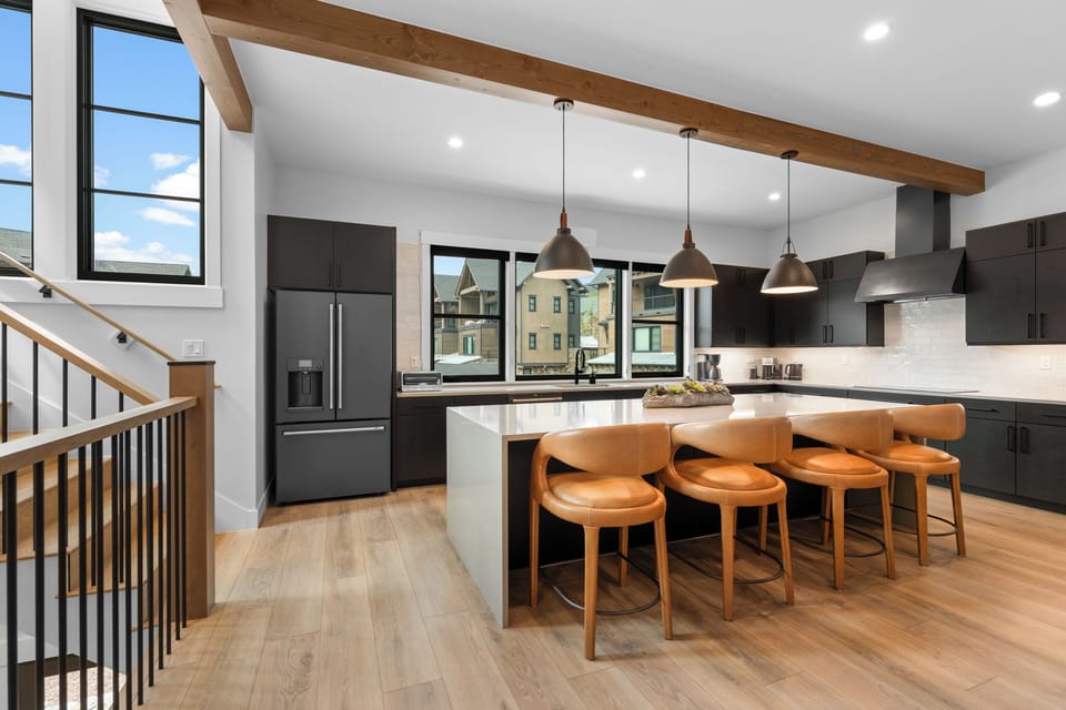 Your modern chef's kitchen features sleek appliances, a spacious island with leather seating for six, and natural light streaming through large windows. The exposed wood beams add warmth to this inviting culinary space.