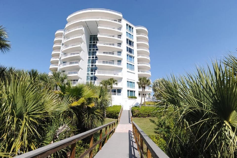 Concrete walkway lined with wooden railings leads through palm trees to a white multi-story condo building.