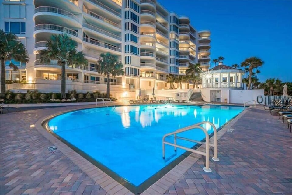 Outdoor pool illuminated at dusk, surrounded by brick pavers, lounge chairs, and palm trees.