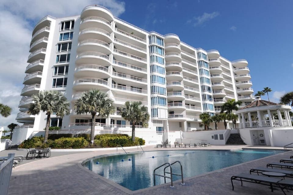 Curved white balconies rise above the kidney-shaped pool with brick pavers and scattered lounge chairs.