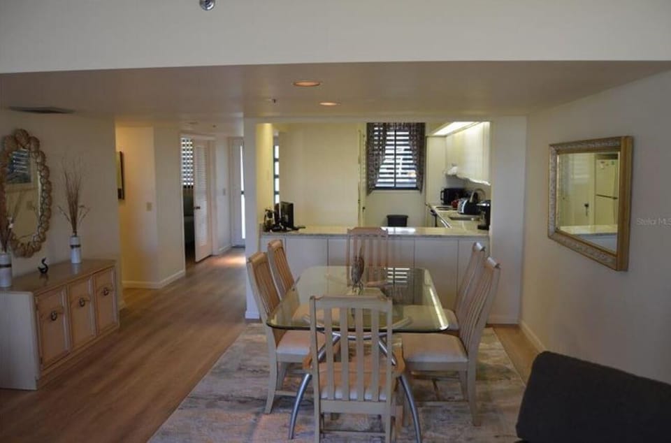 Dining area with glass-top table, six wooden chairs, light wood floors, and a large wall mirror.