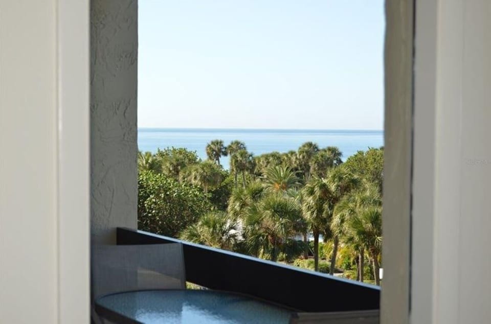 View through living room doorway to balcony with glass-top table and gray mesh chair overlooking palm trees and ocean.