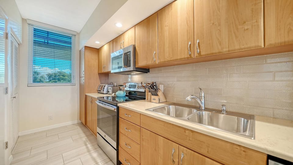 Galley kitchen with light wood cabinets, beige tile backsplash, stainless steel appliances, and a window with blue blinds.