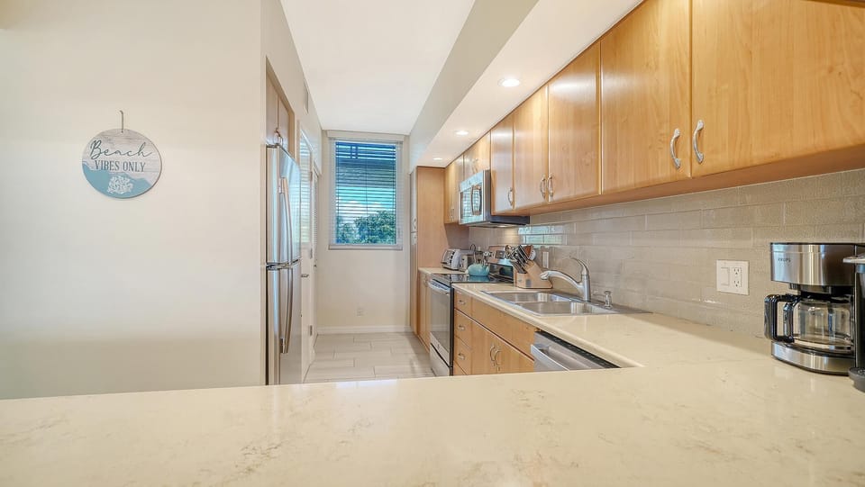 Kitchen with light wood cabinets, quartz countertops, stainless steel appliances, and a window with blinds.