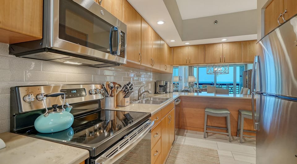 Galley kitchen with light wood cabinets, stone countertops, stainless steel appliances, and a breakfast bar with stools.