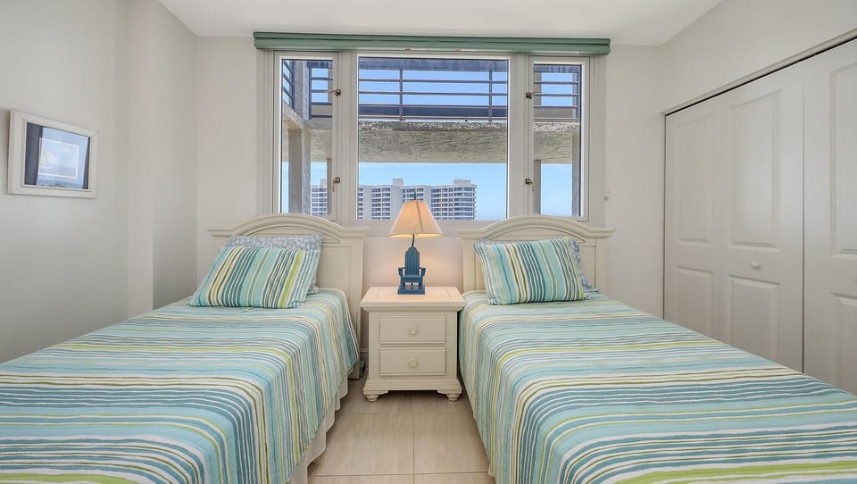 Twin beds with striped blue and green bedding flank a white nightstand topped by a blue lamp beneath a window.