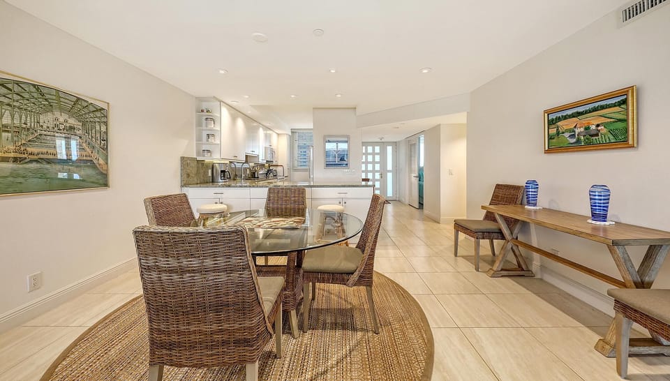 Wicker chairs surround a round glass dining table on a braided jute rug, with tiled floors and artwork on walls.