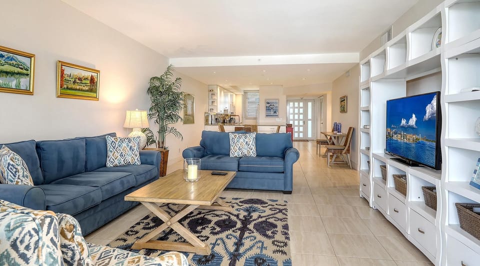 The spacious living room shows blue sofas, patterned armchair, wooden coffee table, and white built-in shelving with TV.