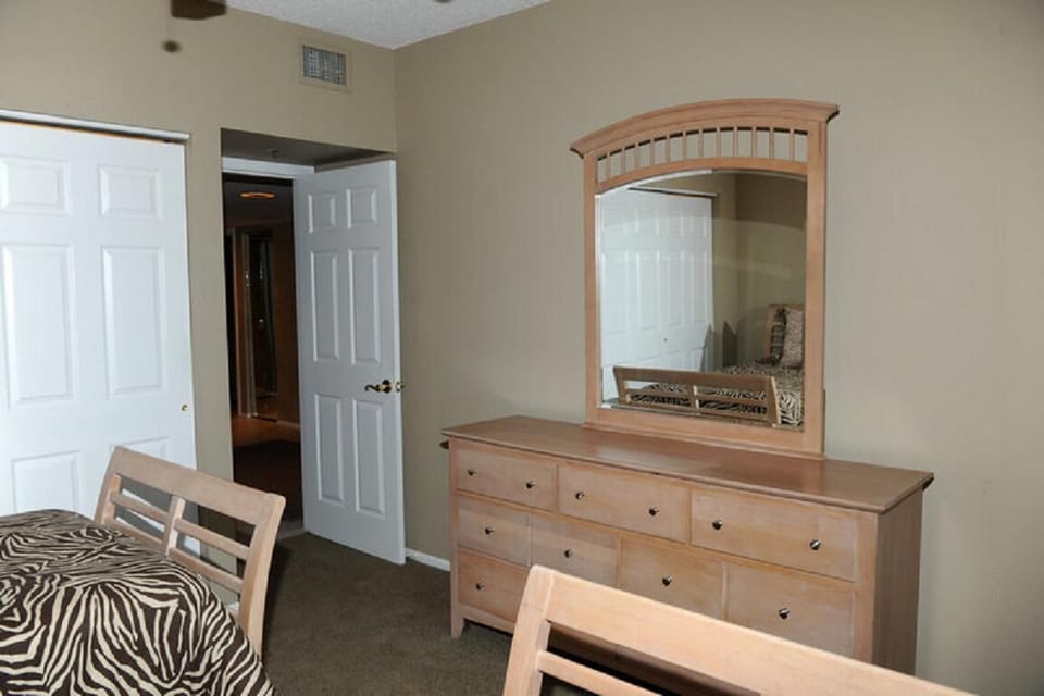 Beige walls and brown carpet frame the bedroom’s wooden bed and dresser with large attached mirror.