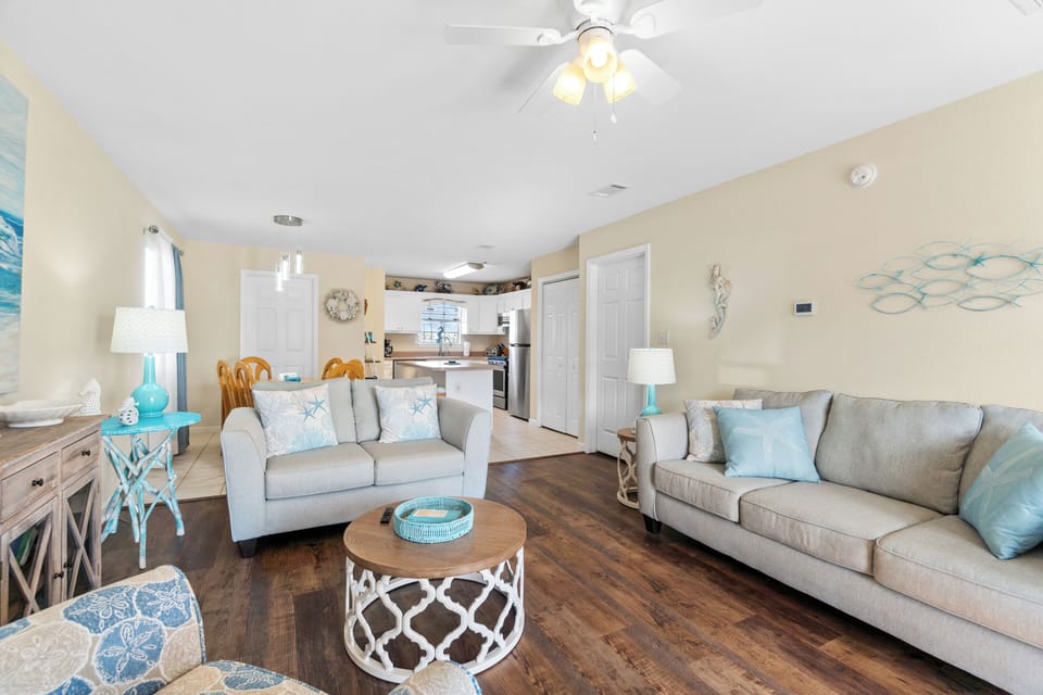 Relax in your coastal-themed living space with soft gray sofas, ocean-blue accents, and an open kitchen perfect for your beach getaway.