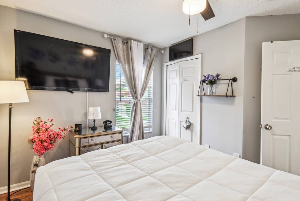 Enjoy comfort in this inviting bedroom with a plush white bedspread and a wall-mounted TV for entertainment. Natural light pours through chic curtains, highlighting the soothing gray tones and cozy decor for a relaxing atmosphere.