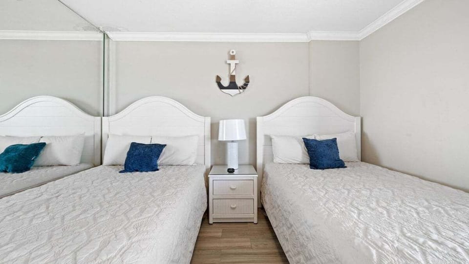 Charming guest room with two cozy twin beds, crisp white linens, and nautical accents. Shared nightstand with modern lamp adds function and style.